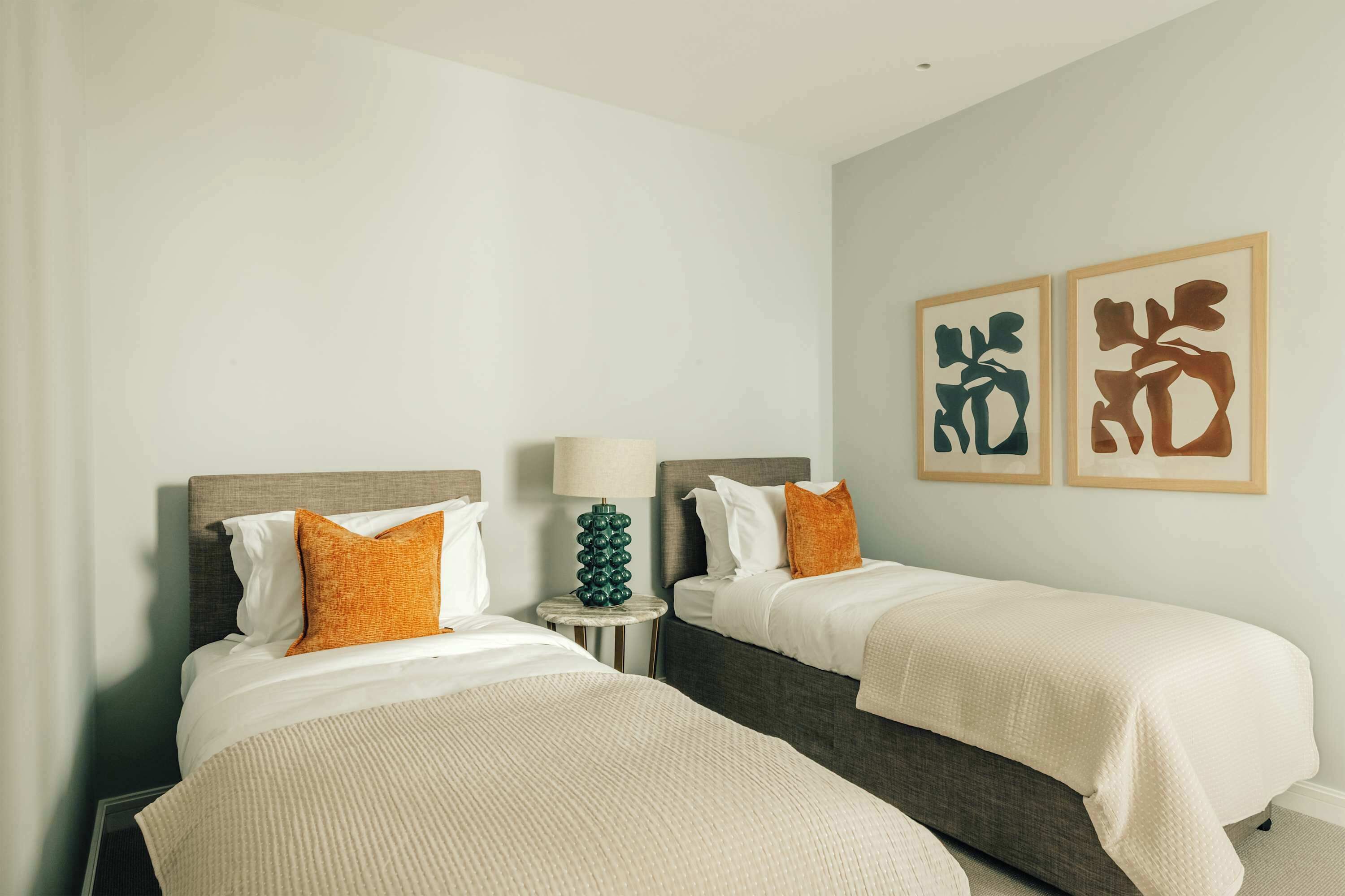 Een hotelkamer met twee bedden, een nachtkastje met lamp en twee abstracte kunstwerken in lijsten aan de muur.