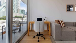 Une pièce moderne avec une grande fenêtre, un bureau, un fauteuil de bureau gris, un canapé gris et deux tableaux encadrés au mur.
