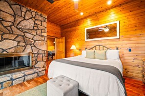 Une chambre avec une cheminée en pierre, des murs en bois, un lit avec des oreillers, une table de chevet et un ventilateur de plafond.