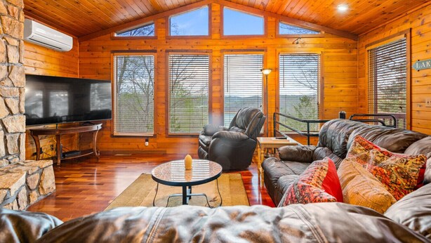 L’intérieur d’une cabane en rondins, avec une cheminée en pierre, un téléviseur, des meubles en cuir et de grandes fenêtres.