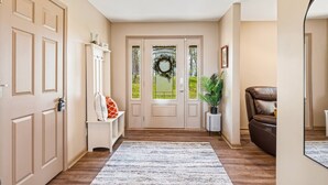 A hallway with a wooden floor, a white rug, a door with a wreath, a bench with cushions, a potted plant, and a leather chair. - The Hideaway: Large (7BD), Modern, Lakeview, Slip (LaFollette)