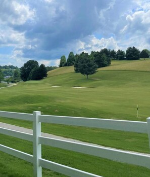 A golf course with a white fence, green grass, and trees in the background.