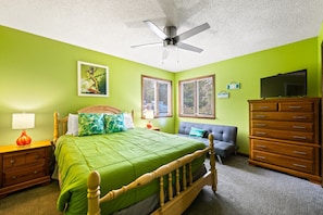 A bedroom with a bed, a dresser, a nightstand, a lamp, a ceiling fan, and a window.