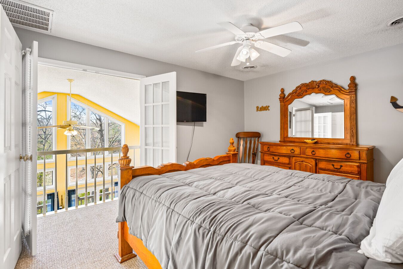 A bedroom with a bed, a dresser, a ceiling fan, a TV, and a balcony with a yellow railing.