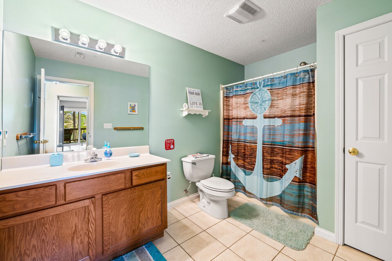 A bathroom with a wooden vanity, a white toilet, a shower curtain with an anchor design, and a green rug.