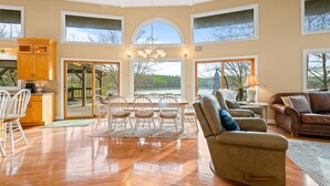 A spacious living area with a wooden floor, a dining table with chairs, a leather sofa, and a view of a lake through large windows. - Poolside Paradise: Lakefront; Private Pool; Dock (LaFollette)