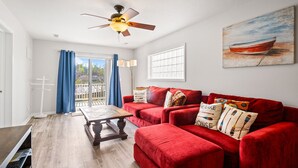 A living room with a red sofa, a wooden coffee table, a ceiling fan, and a view of a balcony with blue curtains.