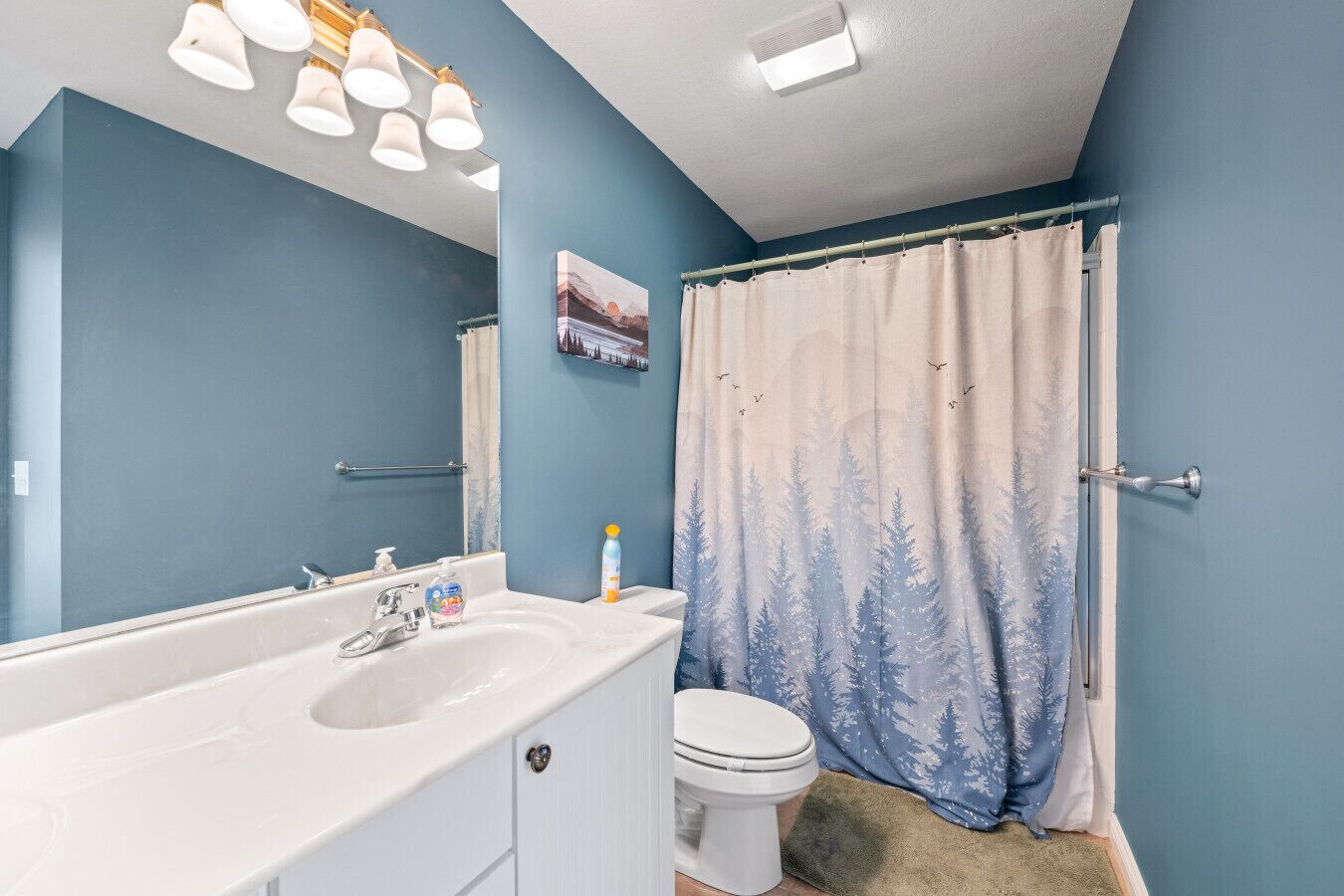 A bathroom with a white sink, a toilet, a shower curtain with a forest pattern, and a mirror with lights above.