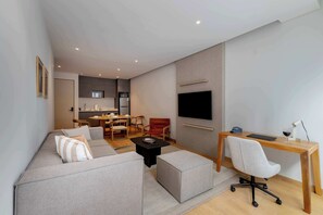 Een moderne woonkamer met een bank, een poef en een houten bureau met een stoel. Er is een flatscreen televisie aan de muur en een eethoek op de achtergrond.