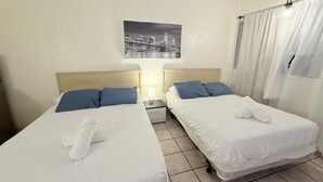 Une chambre d’hôtel avec deux lits, chacun agrémenté de draps blancs et d’oreillers bleus, une table de chevet avec une lampe, et un tableau représentant un paysage urbain accroché au mur.