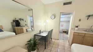 Une chambre d’hôtel de taille réduite, comprenant un lit, un petit bureau, une chaise, une plante en pot, un miroir, une horloge et une kitchenette.