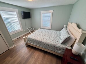 Ein Schlafzimmer mit einem Bett, einem an der Wand montierten Fernseher und zwei Fenstern mit Jalousien.