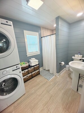 A laundry area with a front-loading washer and dryer, a window with blinds, a shower curtain, a sink, and a small trash bin.