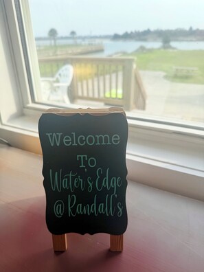 A small black sign with teal text reading 'Welcome To Water's Edge @ Randall's' placed on a wooden stand by a window overlooking a body of water.