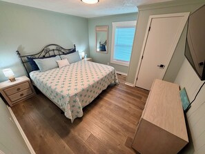 A bedroom with a bed, nightstand, dresser, and a window with blinds. - Water’s Edge at Randall’s - Cottage Overlooking the Intracoastal Waterway (Shallotte)