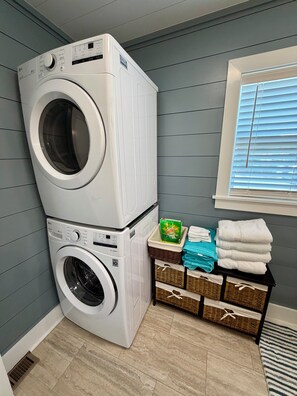 A laundry room with stacked washing machines, baskets with folded towels, and a window with blinds. - Water’s Edge at Randall’s - Cottage Overlooking the Intracoastal Waterway (Shallotte)
