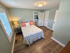 A bedroom with a bed, a nightstand, a lamp, and a window with blinds. - Water’s Edge at Randall’s - Cottage Overlooking the Intracoastal Waterway (Shallotte)