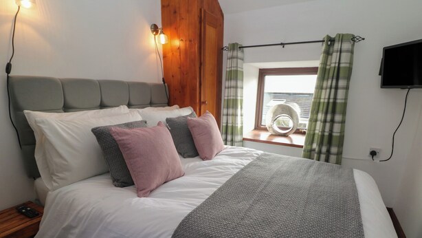 A bedroom with a bed, pillows, a bedside table, a lamp, a window with curtains, and a television.
