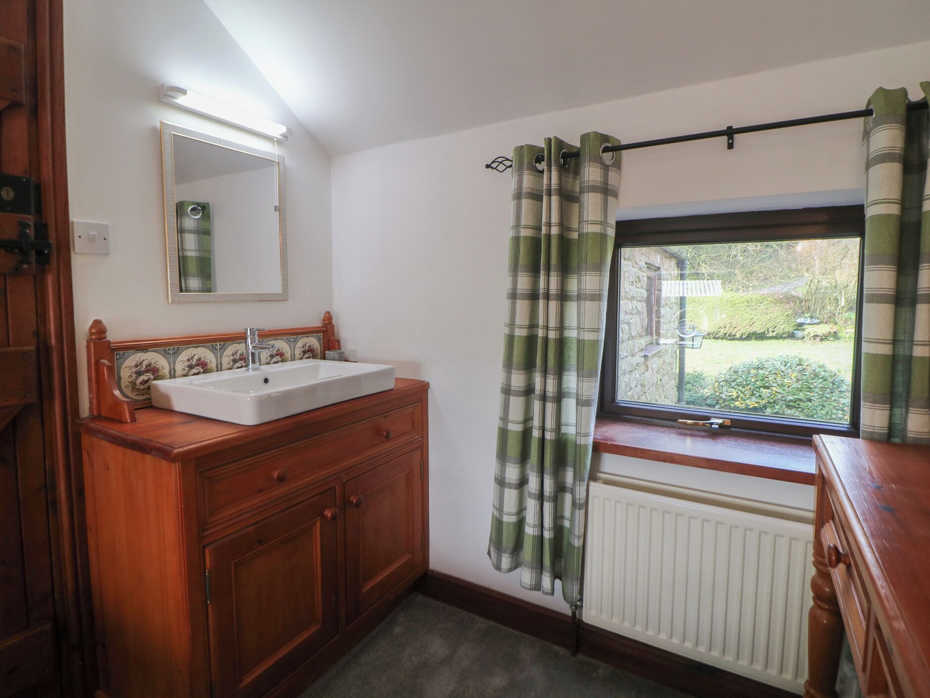 Un baño con tocador de madera, un lavabo blanco, un espejo, una ventana con vistas a follaje y un radiador.