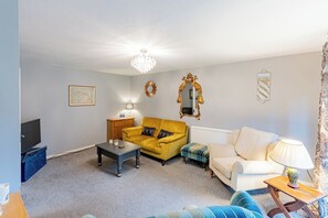 Una sala de estar con un sofá amarillo, un sofá blanco, un sillón azul, una mesa de centro, un televisor y una araña.