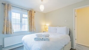 A neatly made bed with white linens and folded towels, a bedside lamp, and a window with curtains.