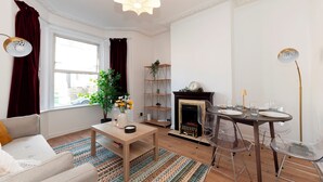 Ein Wohnzimmer mit einer Couch, einem Couchtisch und einem Esstisch für zwei Personen. Ein Kamin und ein Bücherregal sind ebenfalls zu sehen.