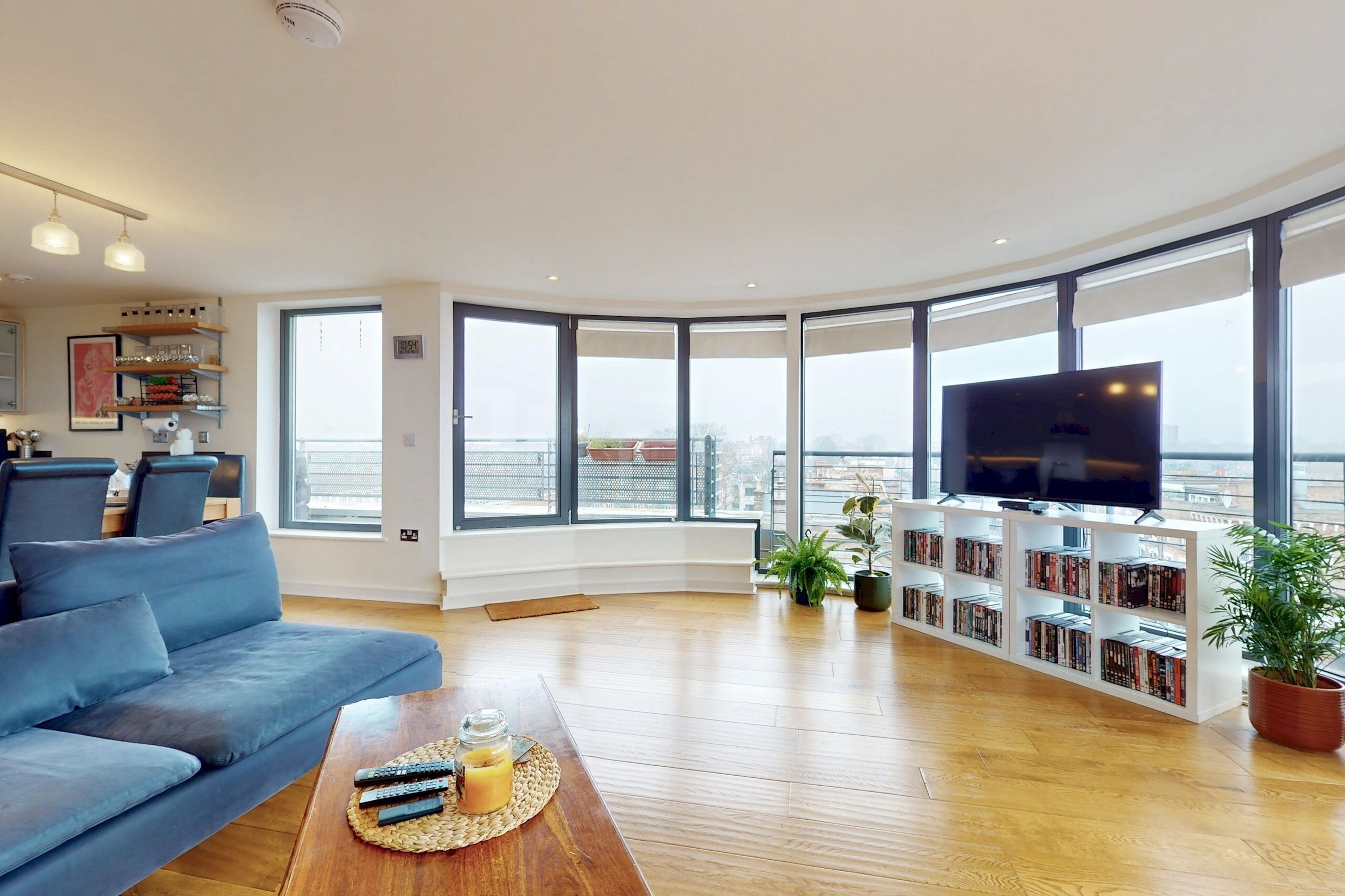 A modern living room with a large flat-screen TV, a blue sofa, a wooden coffee table with remote controls and a drink, and a bookshelf filled with books.