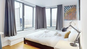 A modern hotel room with a large bed, a desk, a chair, and a window with curtains. - Penthouse Sanctuary with Wraparound Balcony - Pass the Keys (London)