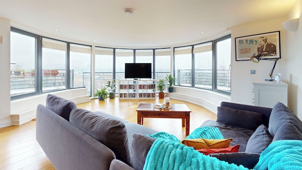 A modern living room with a large TV, a bookshelf, and a view of the city through large windows. - Penthouse Sanctuary with Wraparound Balcony - Pass the Keys (London)