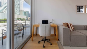 Een moderne kamer met een groot raam, een bureau, een grijze bureaustoel, een grijze bank en twee ingelijste foto's aan de muur.