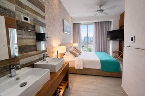 A hotel room with a large bed, a bathroom with a sink and mirror, a ceiling fan, and a TV. - Apartamento Amueblado en Naco – Piso 12 (Santo Domingo)