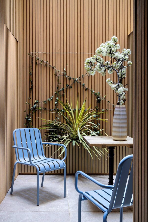 Área de estar com duas cadeiras, uma mesa, planta em vaso e um vaso de flores, tudo em frente a uma parede de madeira.