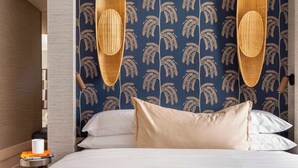 A hotel room with a bed, two wicker chairs, a patterned wallpaper, and a bedside table with a book and a cup. - Luxury Garden Suit in Recoletos (Madrid)