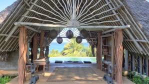 Un pavillon au toit de chaume, agrémenté d’un élément décoratif central, un sentier de pierre menant à une piscine bordée de nénuphars et une vue sur l’océan.