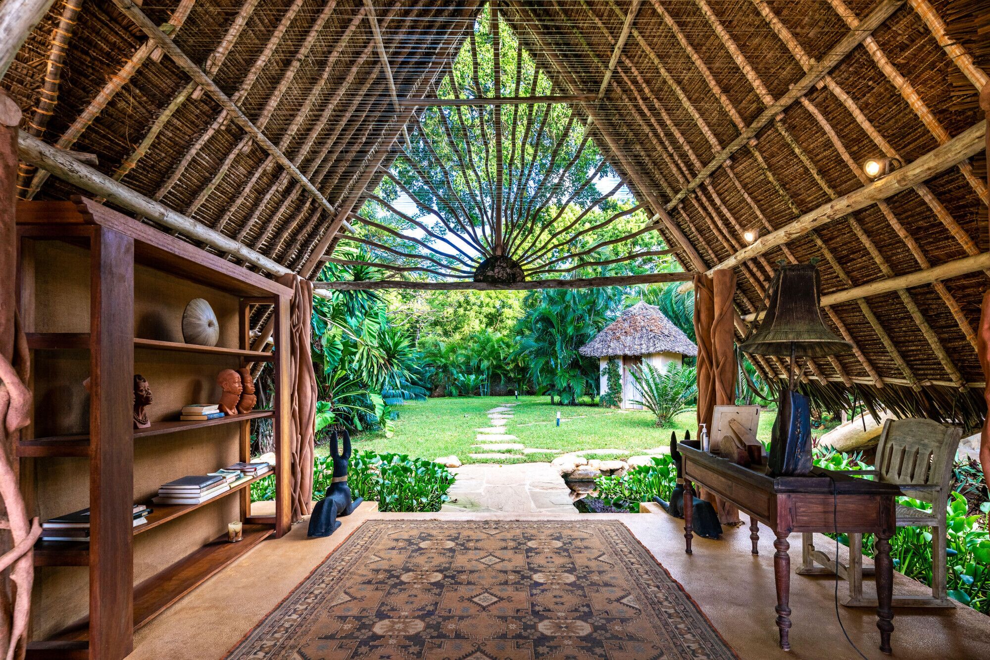 Un interior con techo de paja, estantes de madera, alfombra estampada y vista a un jardín exuberante.