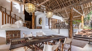 Un coin repas rustique avec une longue table en bois, des chaises rembourrées de couleur blanche et un toit de chaume.