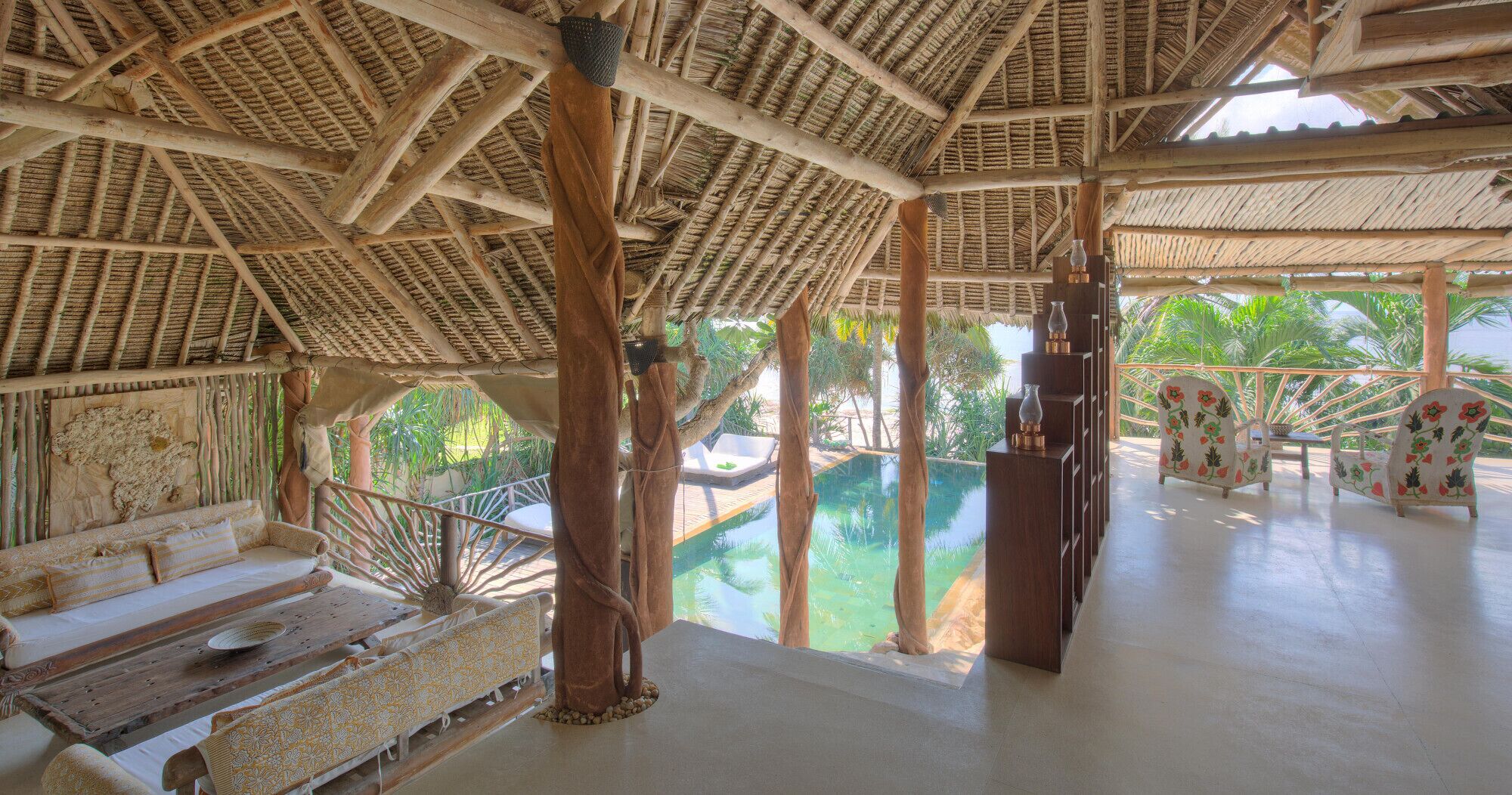 A spacious lounge area with a pool, wooden furniture, and a thatched roof structure.