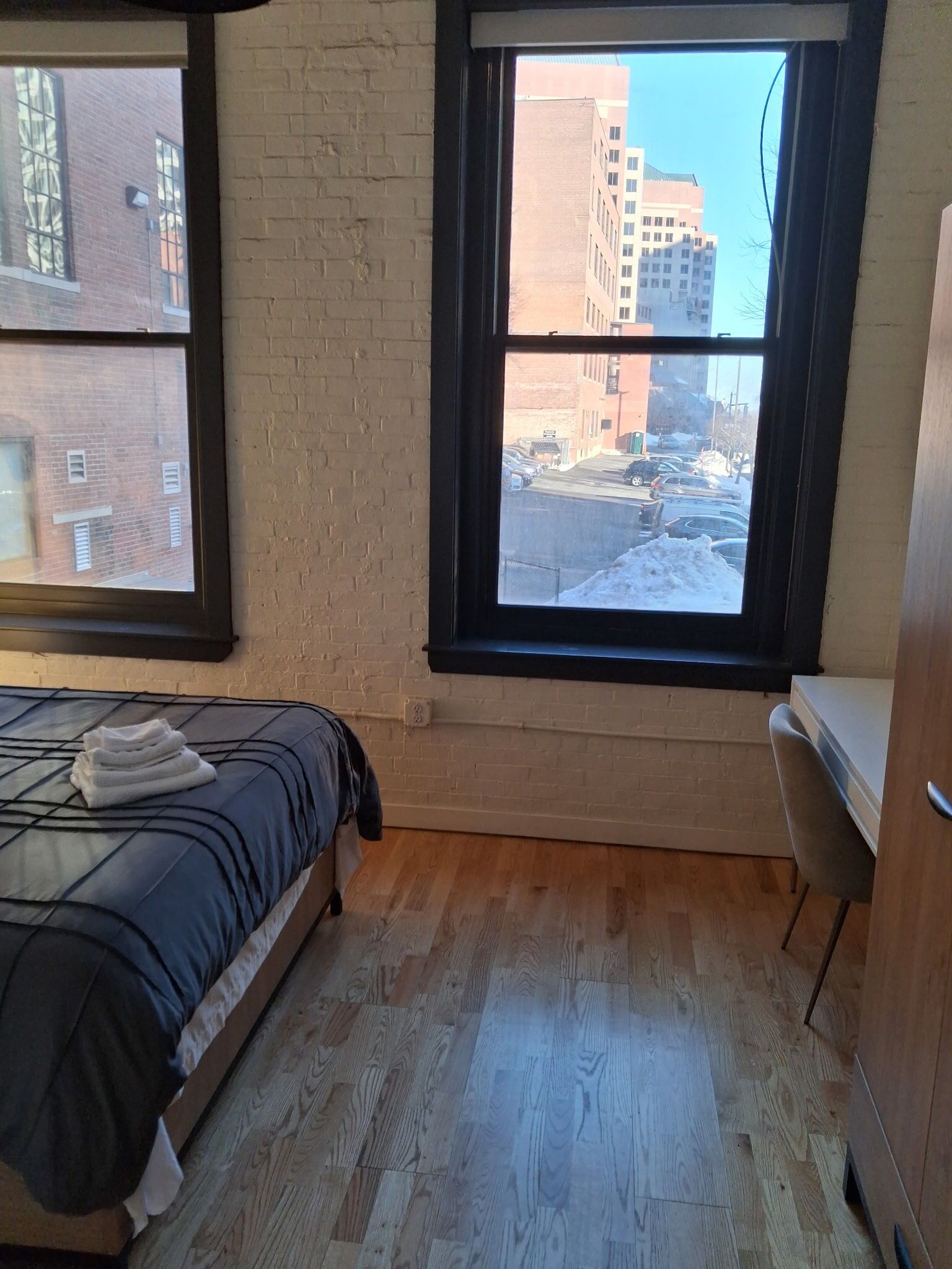 Ein Zimmer mit einem Bett, einem Fenster mit Blick auf Gebäude und einen Parkplatz, sowie einem Holzboden.