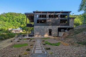 Un bâtiment moderne à deux étages, doté d’une façade en pierre, d’un espace extérieur couvert avec des sièges et d’un chemin en pierre menant à l’entrée.
