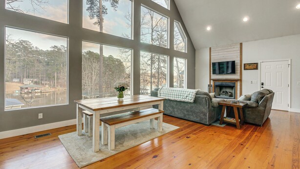 Ein geräumiges Wohnzimmer mit Holztisch, Bank und grauem Sofa, große Fenster mit Blick auf Bäume und einen Steg, sowie ein Kamin.