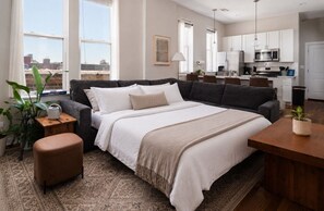 A modern bedroom with a large bed, a small side table, a coffee table, and a view of the city through large windows.