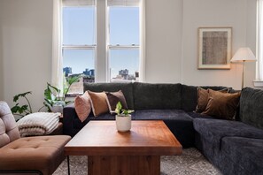 A living room with a dark grey sofa, a wooden coffee table, and a city view through large windows.