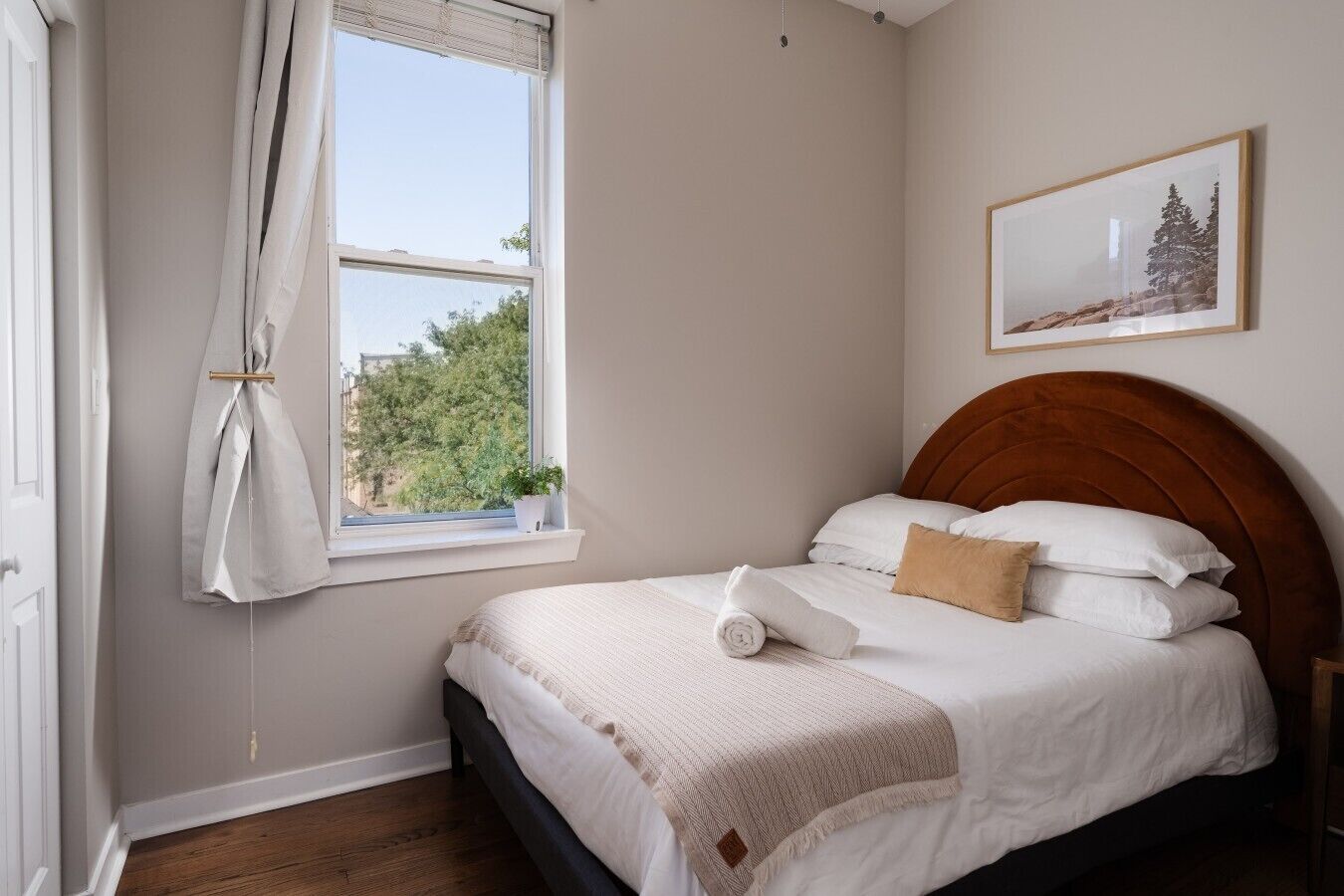 Une chambre à coucher avec un lit, une fenêtre avec des rideaux, un tableau accroché au mur et une tête de lit en bois.