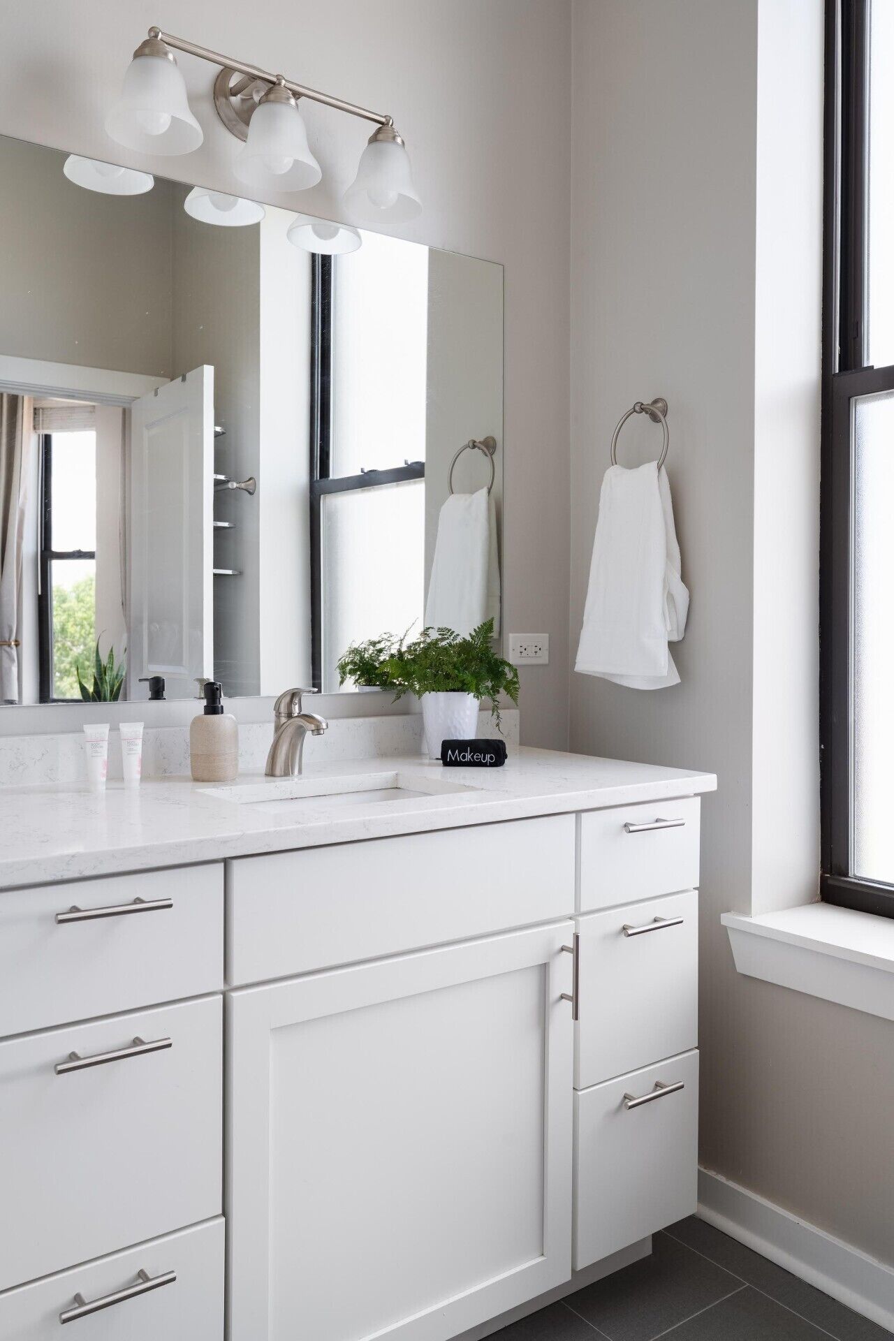A modern bathroom with a white vanity, a large mirror, a light fixture, a potted plant, and a towel rack.