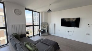 A modern living room with a large window, a sofa, a flat-screen TV, and a radiator. - Sky View Apartment (Barking)