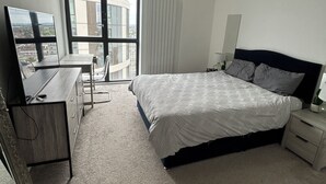 A modern bedroom with a large bed, a desk, and a view of buildings through a large window. - Sky View Apartment (Barking)