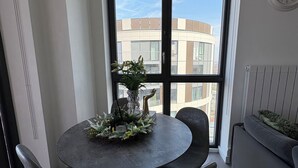 A modern dining area with a round table, four chairs, and a decorative centerpiece. A large window offers a view of a building outside. - Sky View Apartment (Barking)