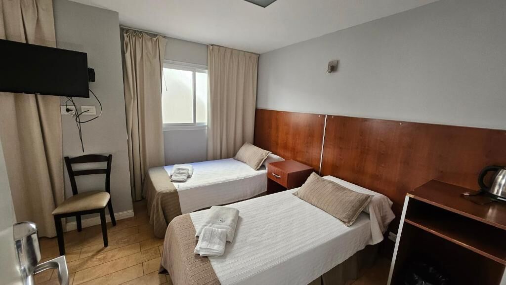 A hotel room with two beds, a television mounted on the wall, a chair, a desk, and a window with curtains.