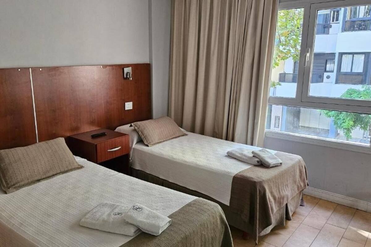 A hotel room with two single beds, wooden headboards, a nightstand, and a window with curtains.