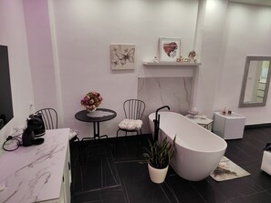 Ein modernes Badezimmer mit einer freistehenden Badewanne, einem kleinen runden Tisch mit Blumen in einer Vase, einem Stuhl und einem Regal mit Dekorationen.